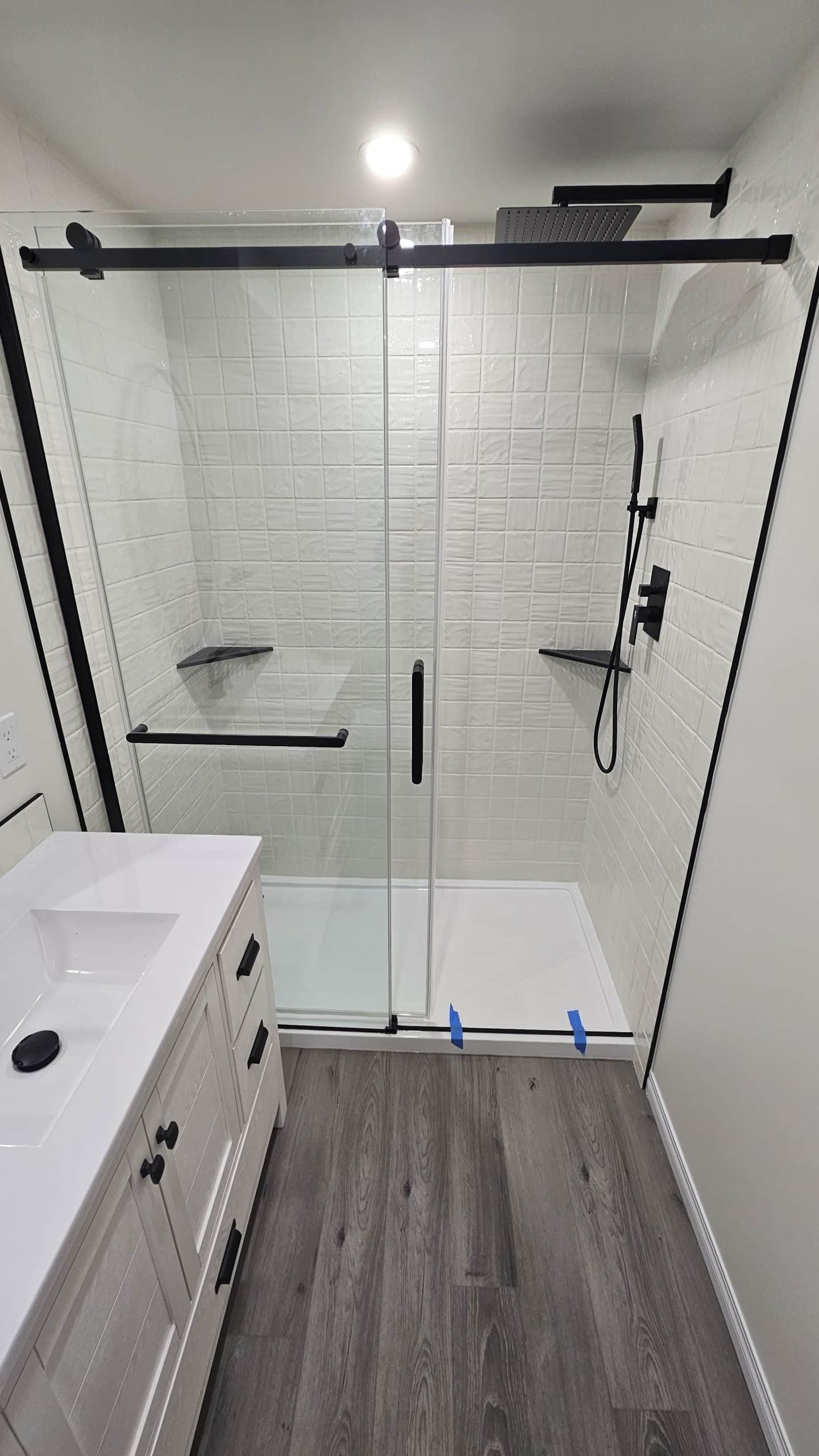 Modern bathroom with a glass shower, black hardware, white vanity, and gray wood floors. Modern bathroom with a glass shower, black hardware, white vanity, and gray wood floors.